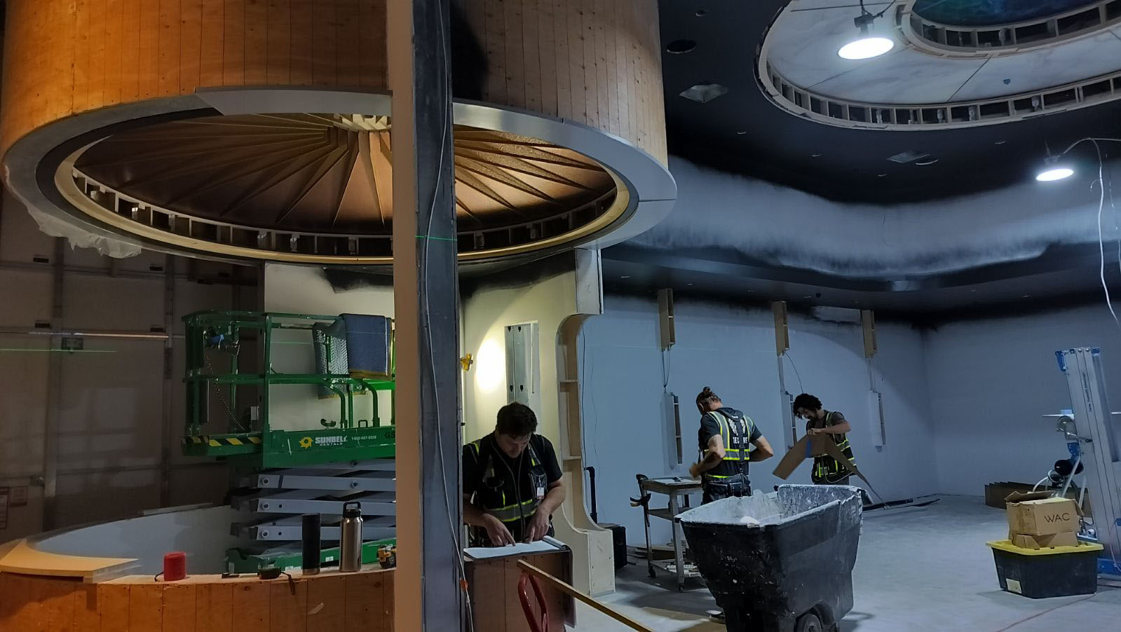 Industrial interior with workers assembling large cylindrical machinery under bright overhead lights.