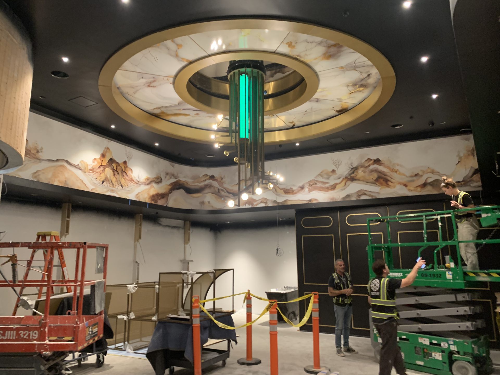 Workers install a large circular ceiling feature with green lighting in a modern lobby or atrium.