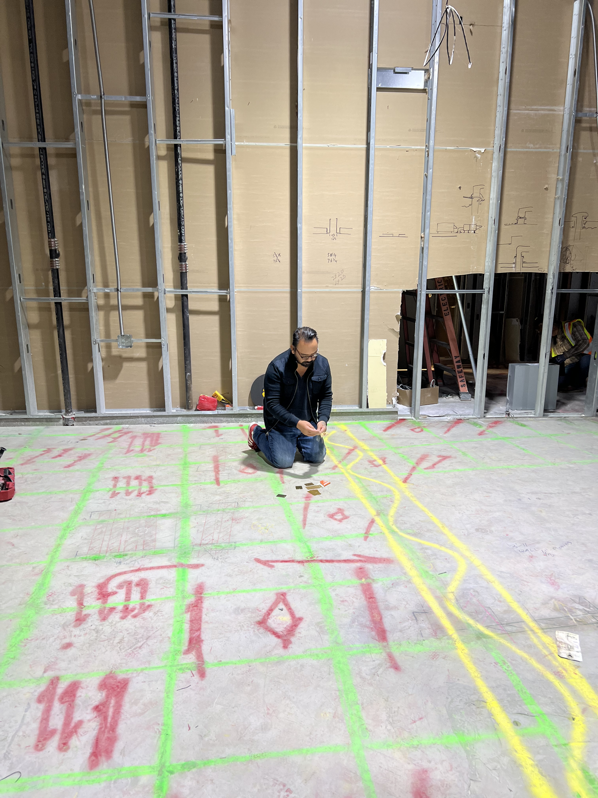 Worker kneeling on marked concrete floor in unfinished room with exposed wall framing and bright yellow cables