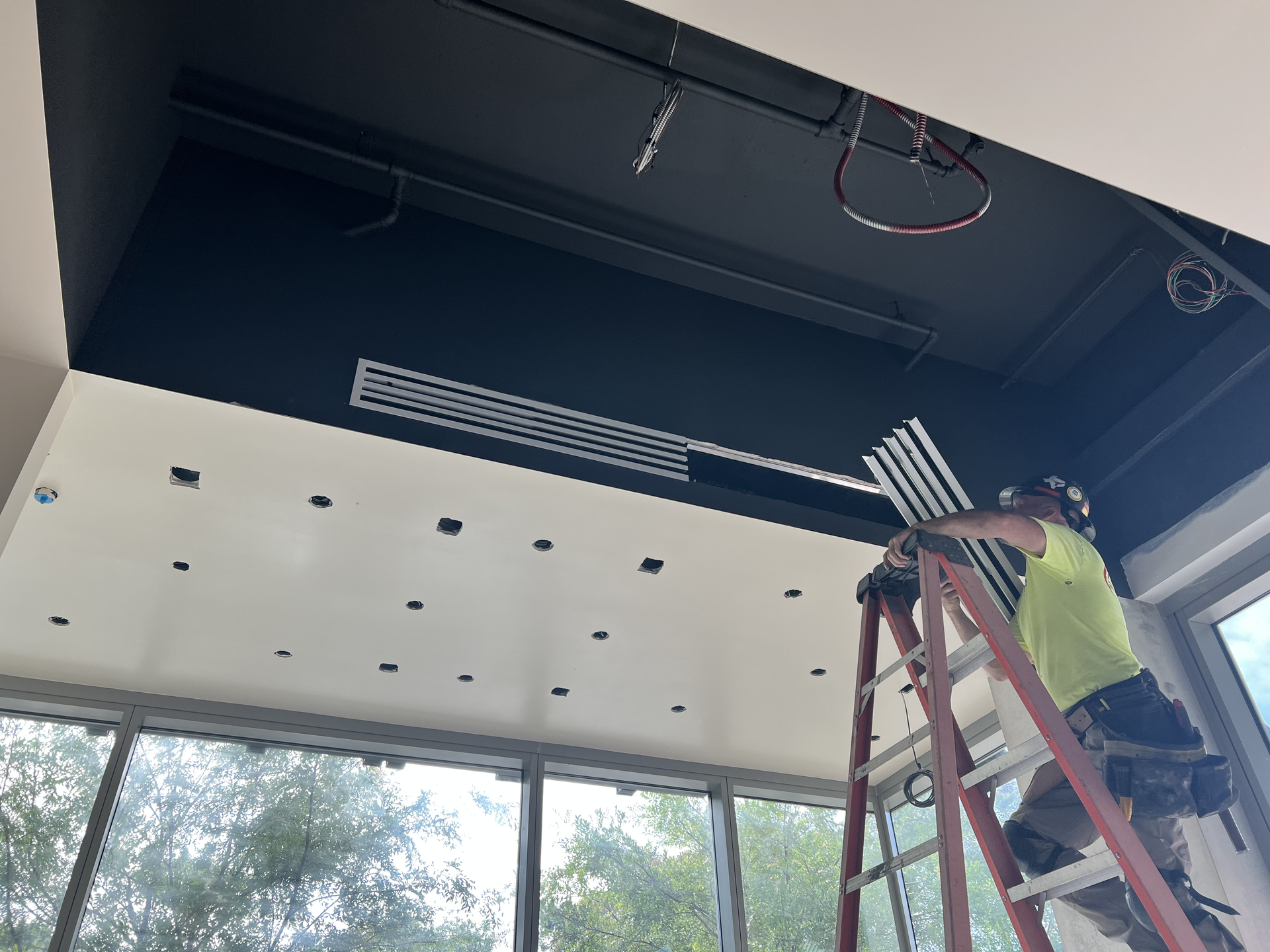 Worker on a ladder installing ceiling fixtures in a modern room with large windows.