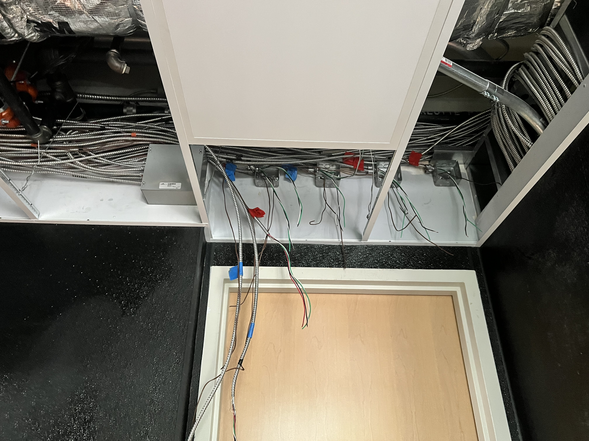 Open ceiling with exposed cables and wiring above a wooden hatch panel in a utility space