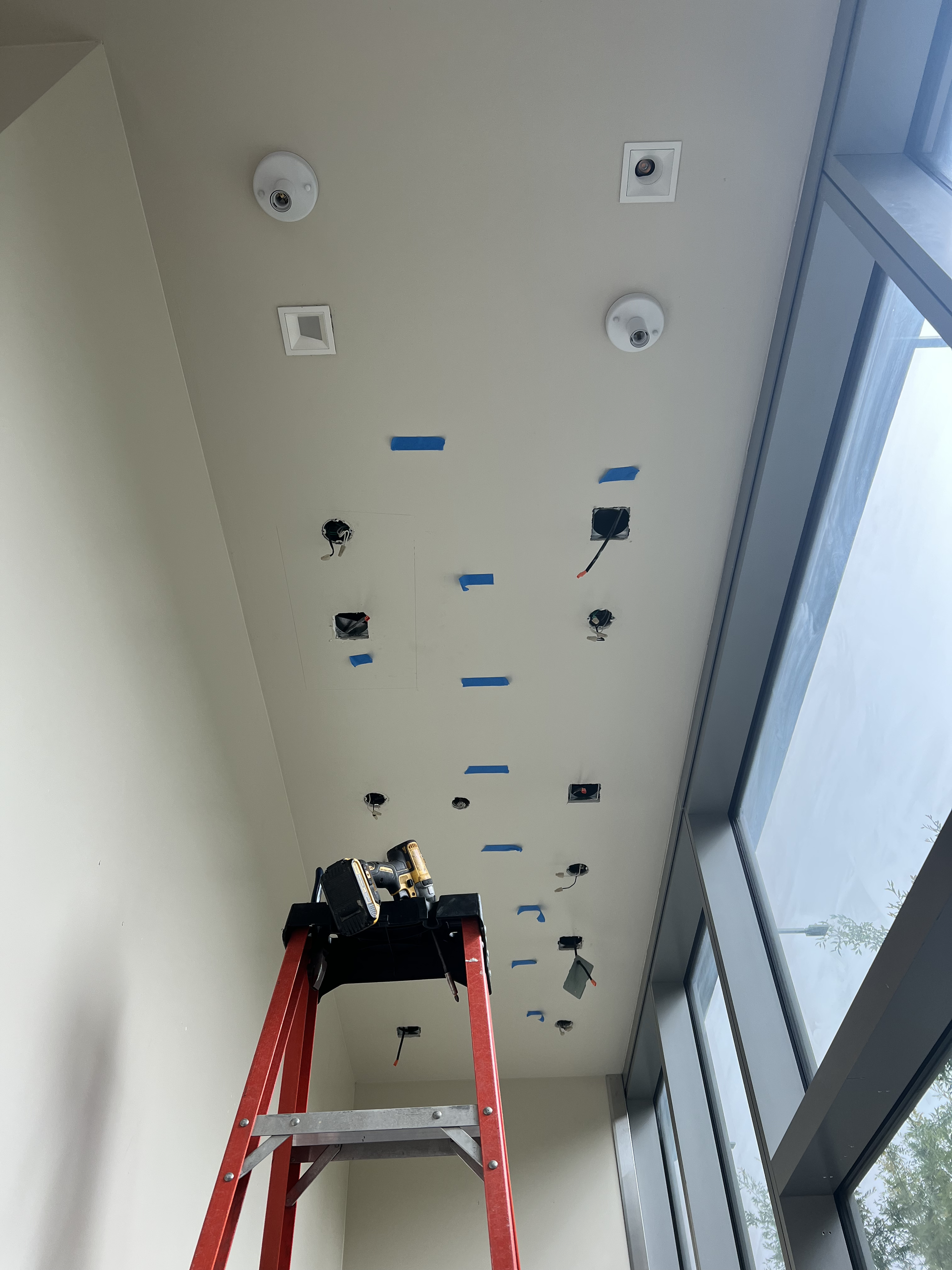 Red ladder beneath a ceiling with smoke detectors and wires in a bright hallway near windows