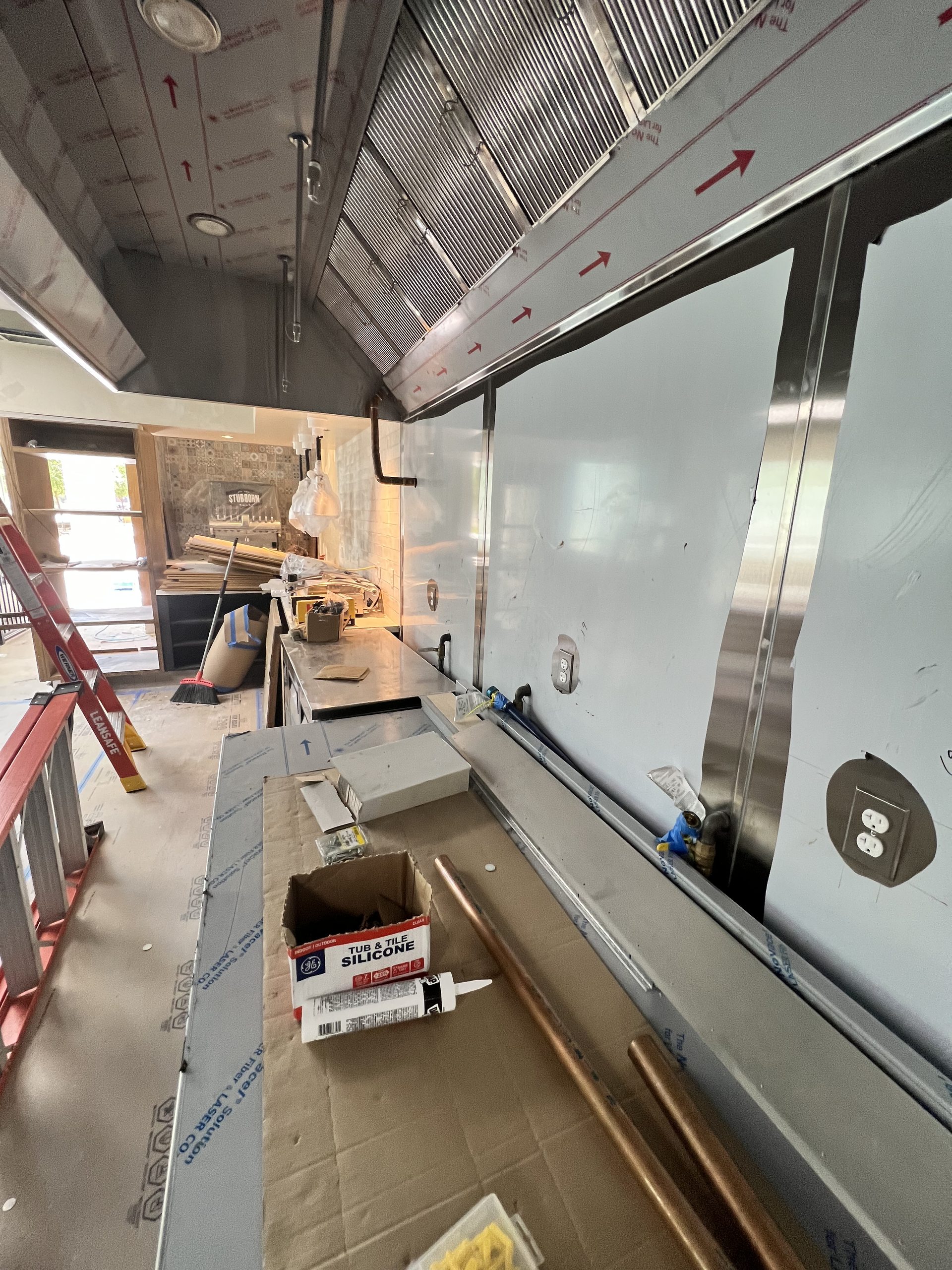 Narrow construction walkway beside a large metal wall, with tools, boxes, and ladders along the corridor
