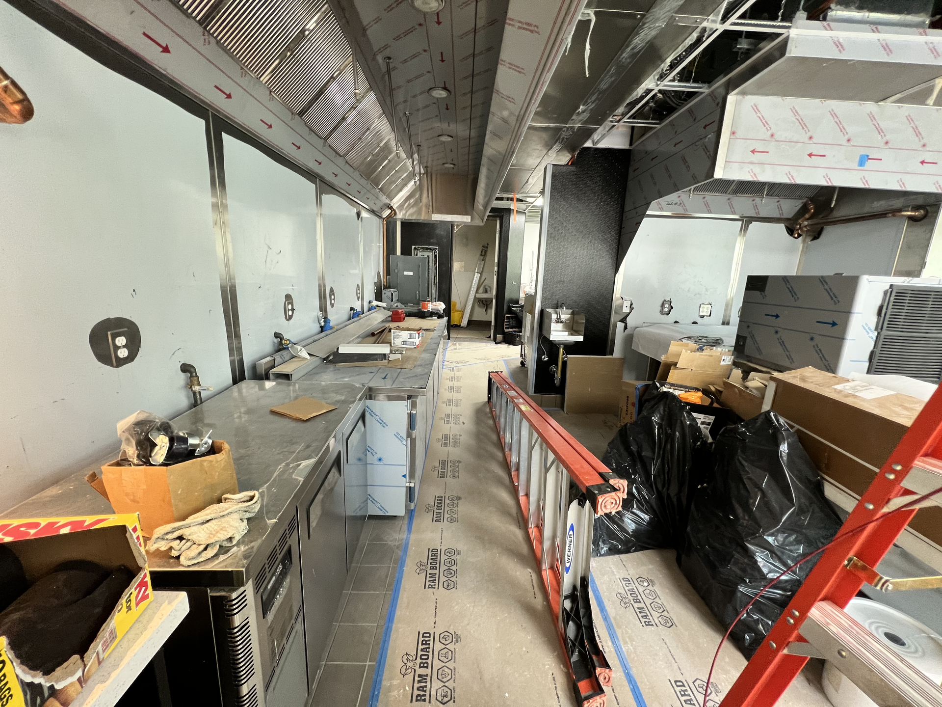 Narrow industrial kitchen under construction with counters, ductwork, boxes, and a red ladder.