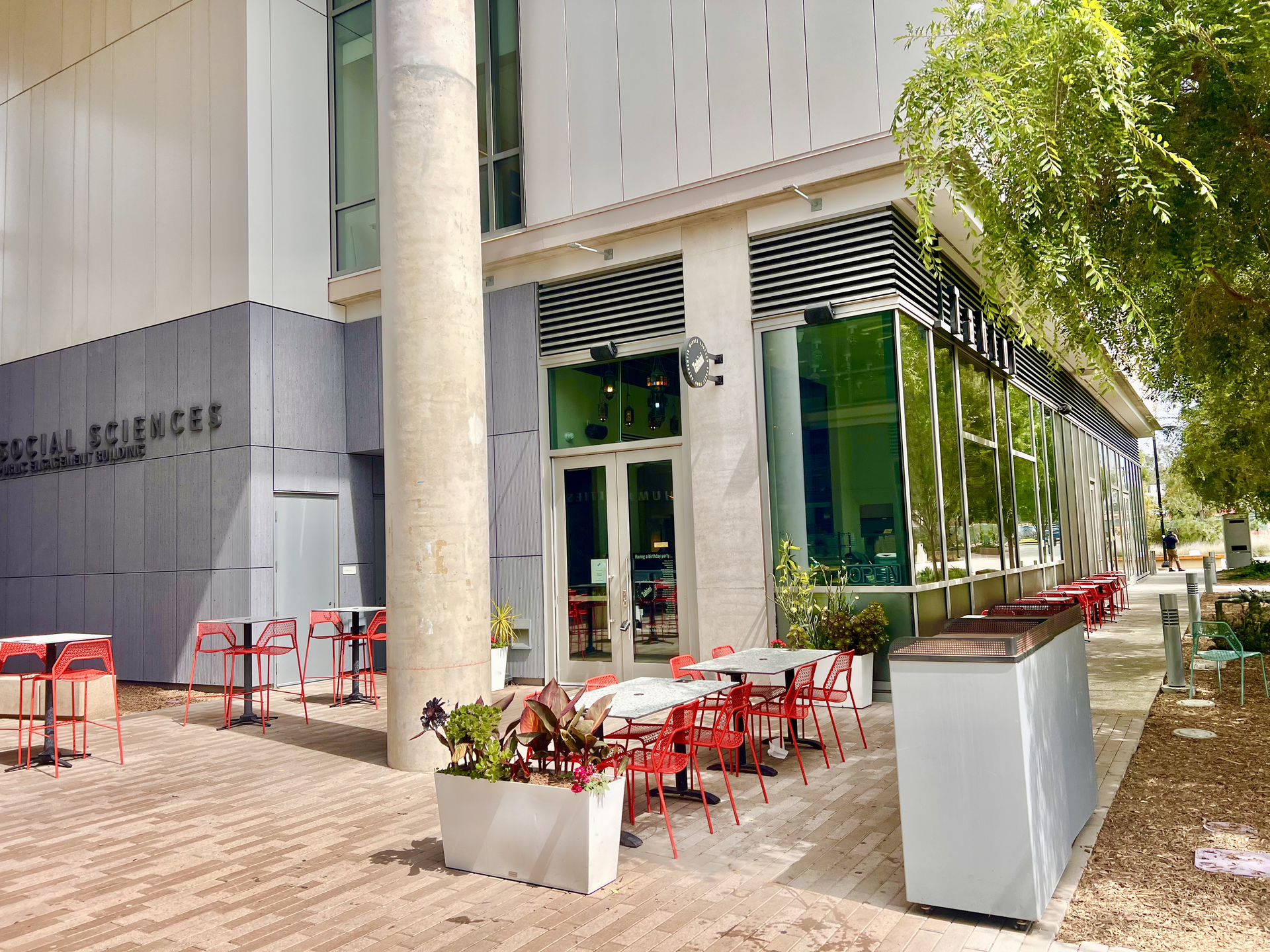Outdoor café with red chairs and tables beside a modern building entrance under a tree shade