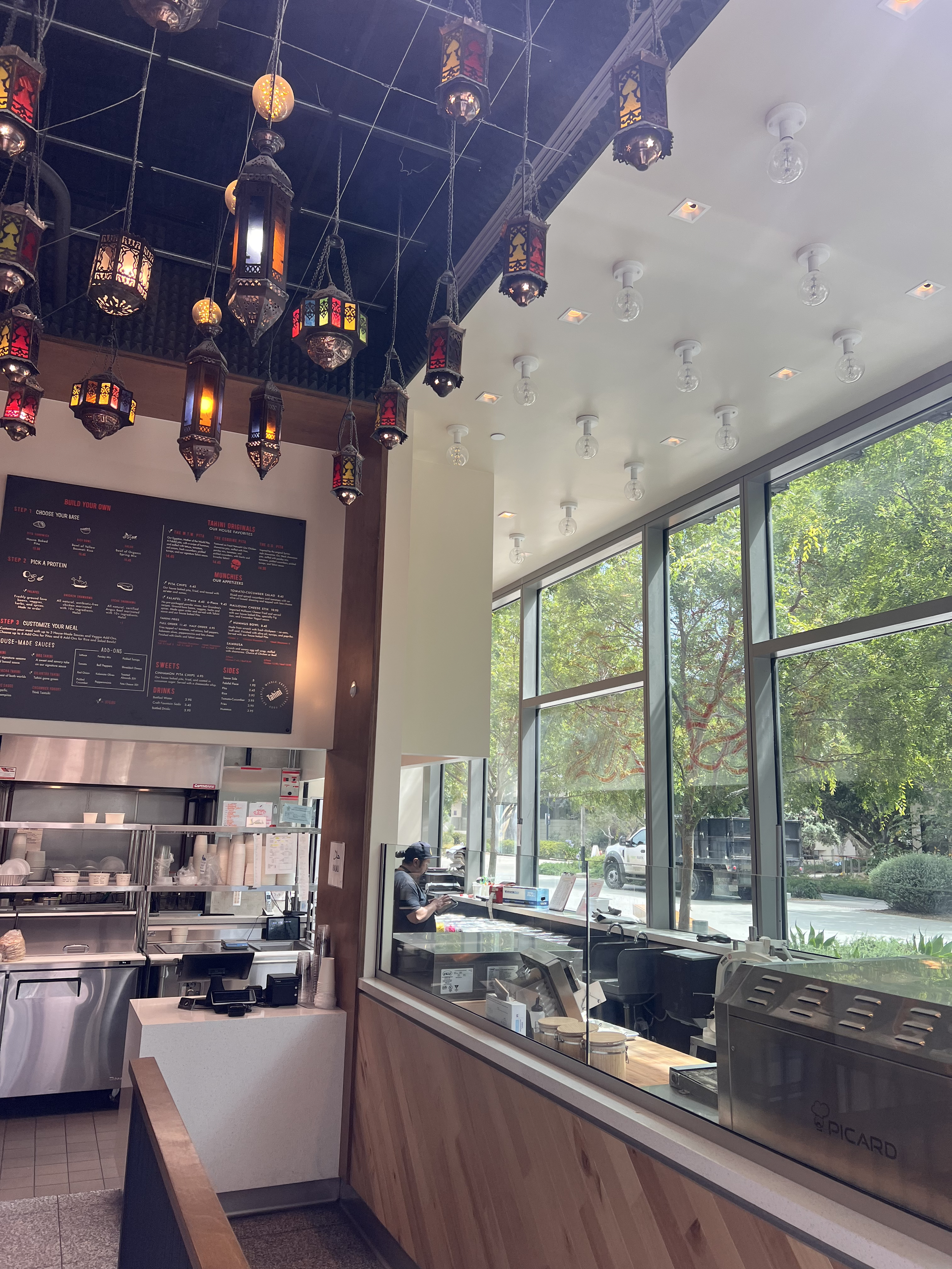 Bright café interior with hanging lanterns, wooden counter, and large windows overlooking greenery.
