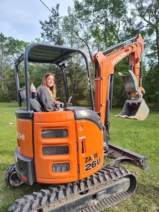 Woman seated on an orange mini excavator on a grassy lawn, with trees in the background
