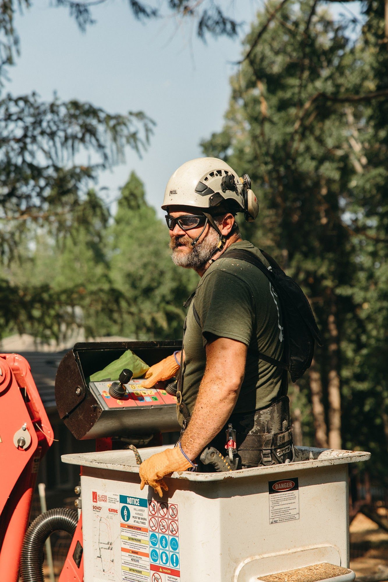 Worker in a hard hat riding in a utility bucket near trees, holding tools and smiling.