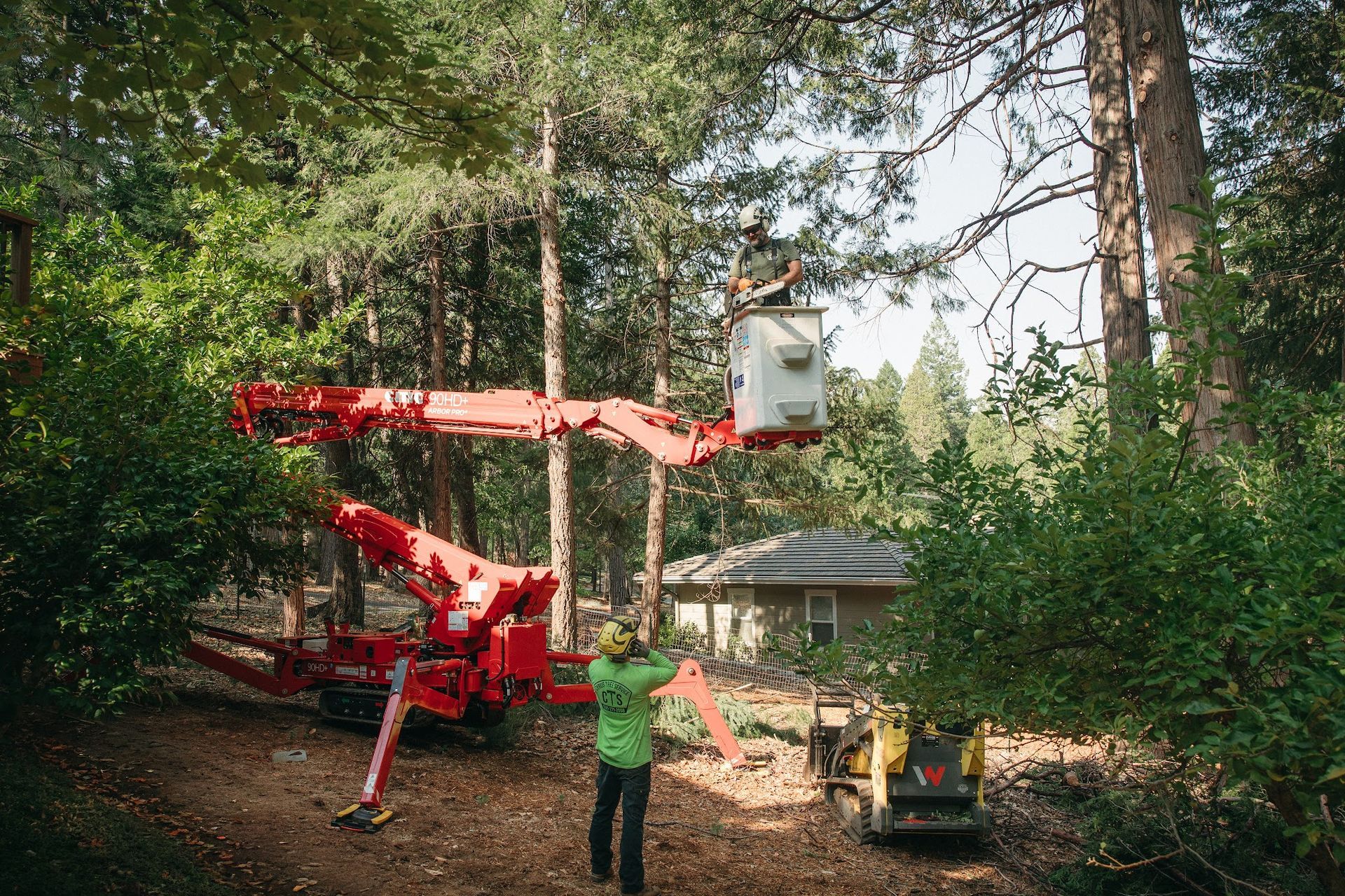 Worker operates a red boom lift in a wooded yard near a house while another worker guides from below