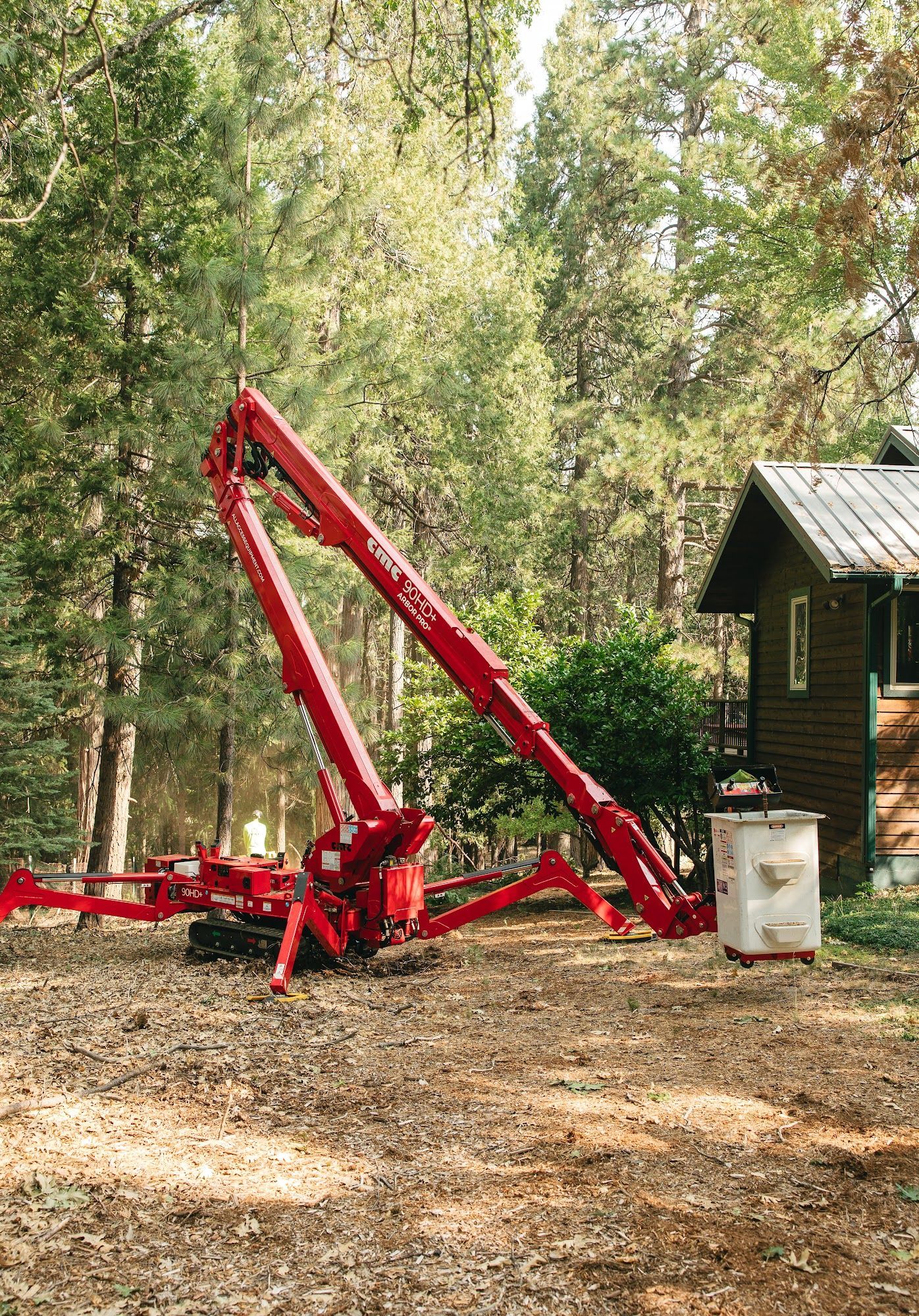 Red forestry grapple loader parked beside a small cabin in a wooded area.