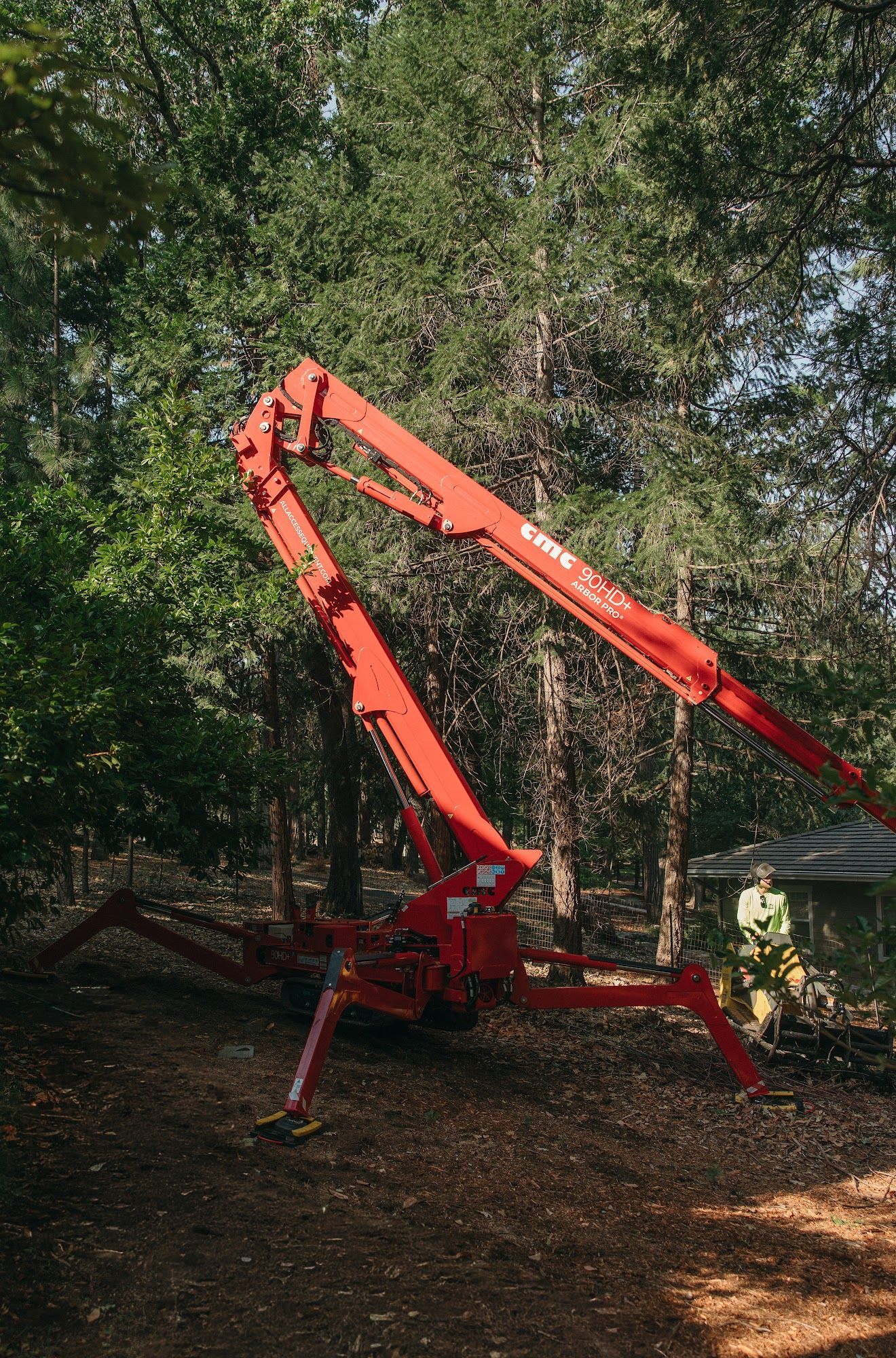Red excavator arm on tripod base in a wooded area, with a person standing nearby