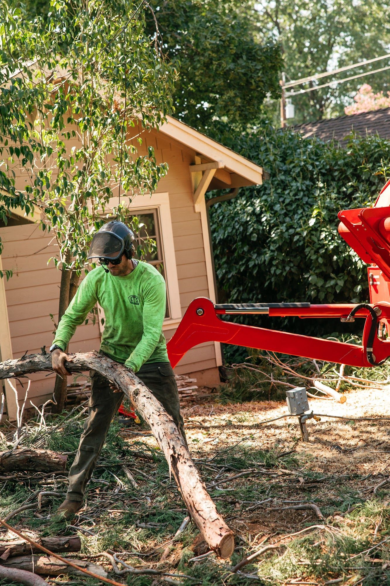 Worker in green shirt cutting a fallen tree branch beside a red excavator in a backyard