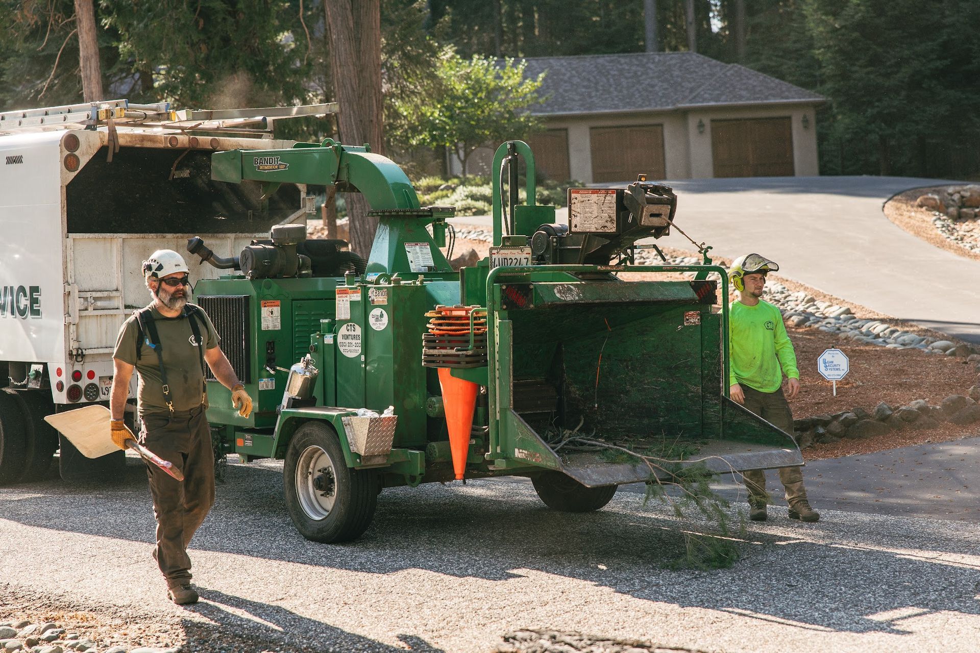 Workers operate a green wood chipper on a residential street, with one worker walking nearby.