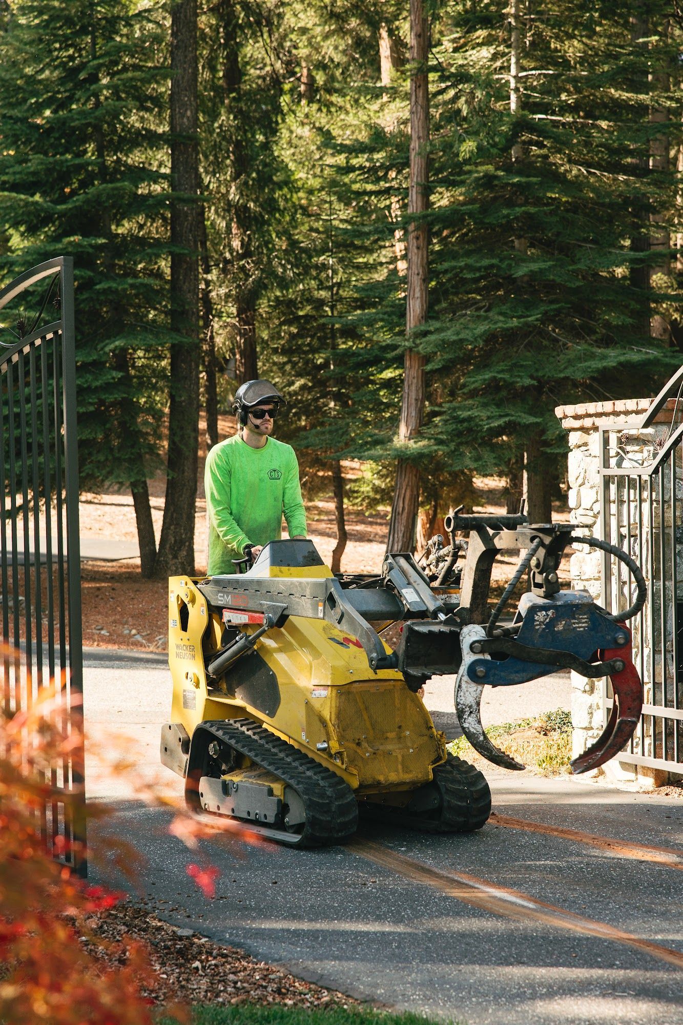 Worker in a neon green shirt operates a yellow tracked machine on a paved path near trees and a fence.