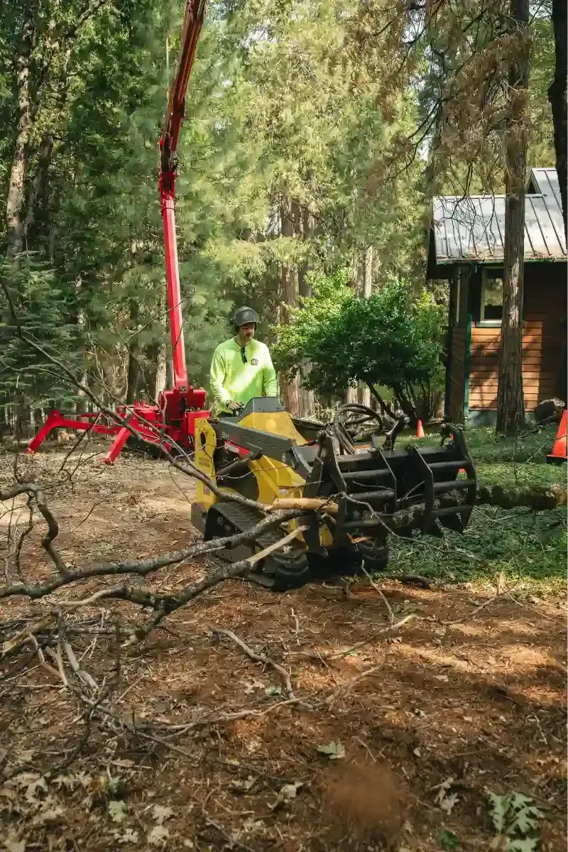 Tree-cutting machine clearing fallen branches in a wooded yard, with a worker in a neon vest nearby.