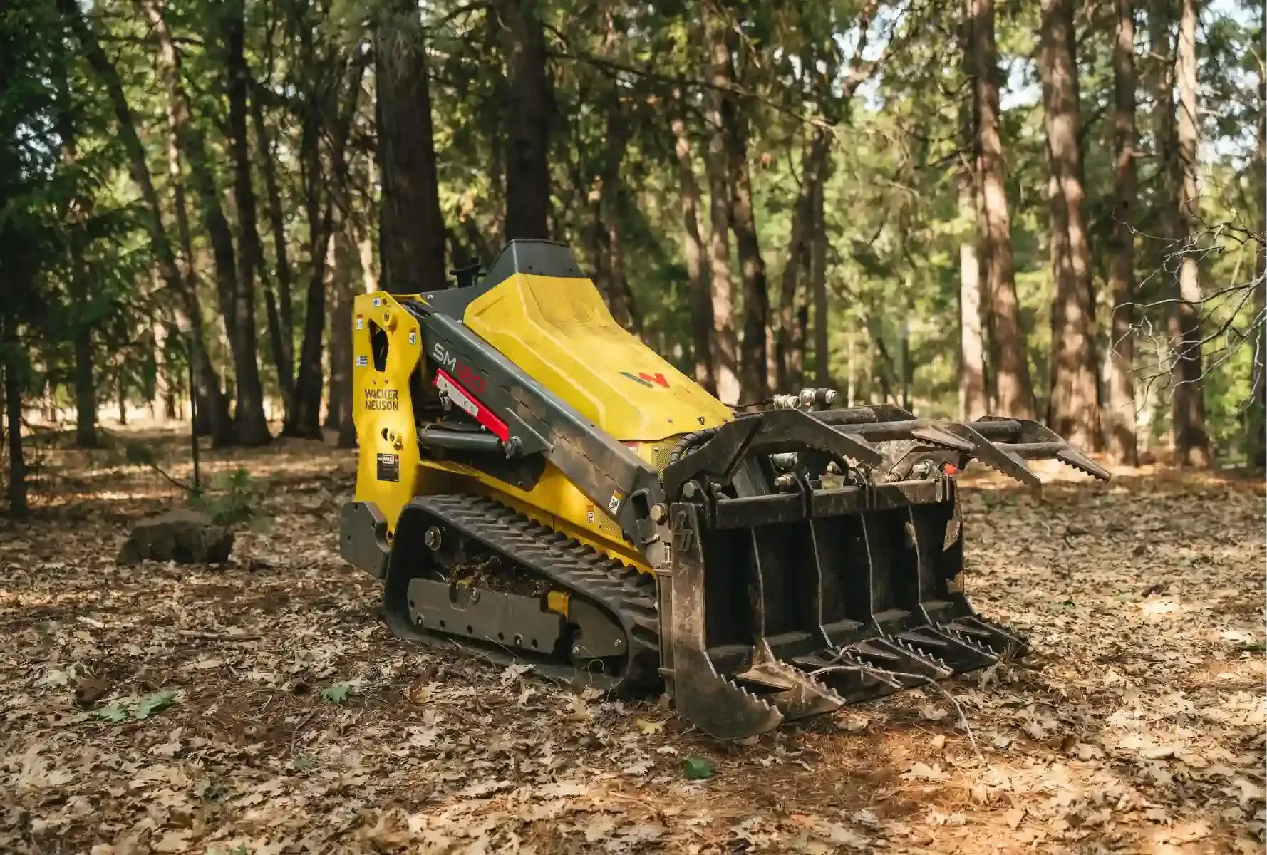 Yellow tracked forestry machine in a wooded area, with a front grapple attachment on leaf-covered ground.