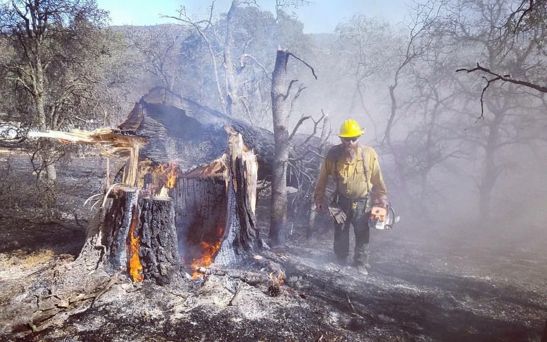 Firefighter walking beside a burning charred structure in a smoky, burned landscape