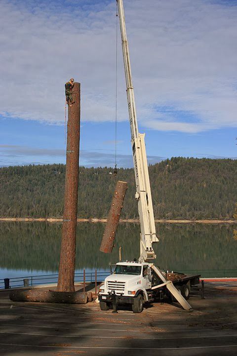 Crane lifting a tall brown piling beside a white truck on a lakeshore work site
