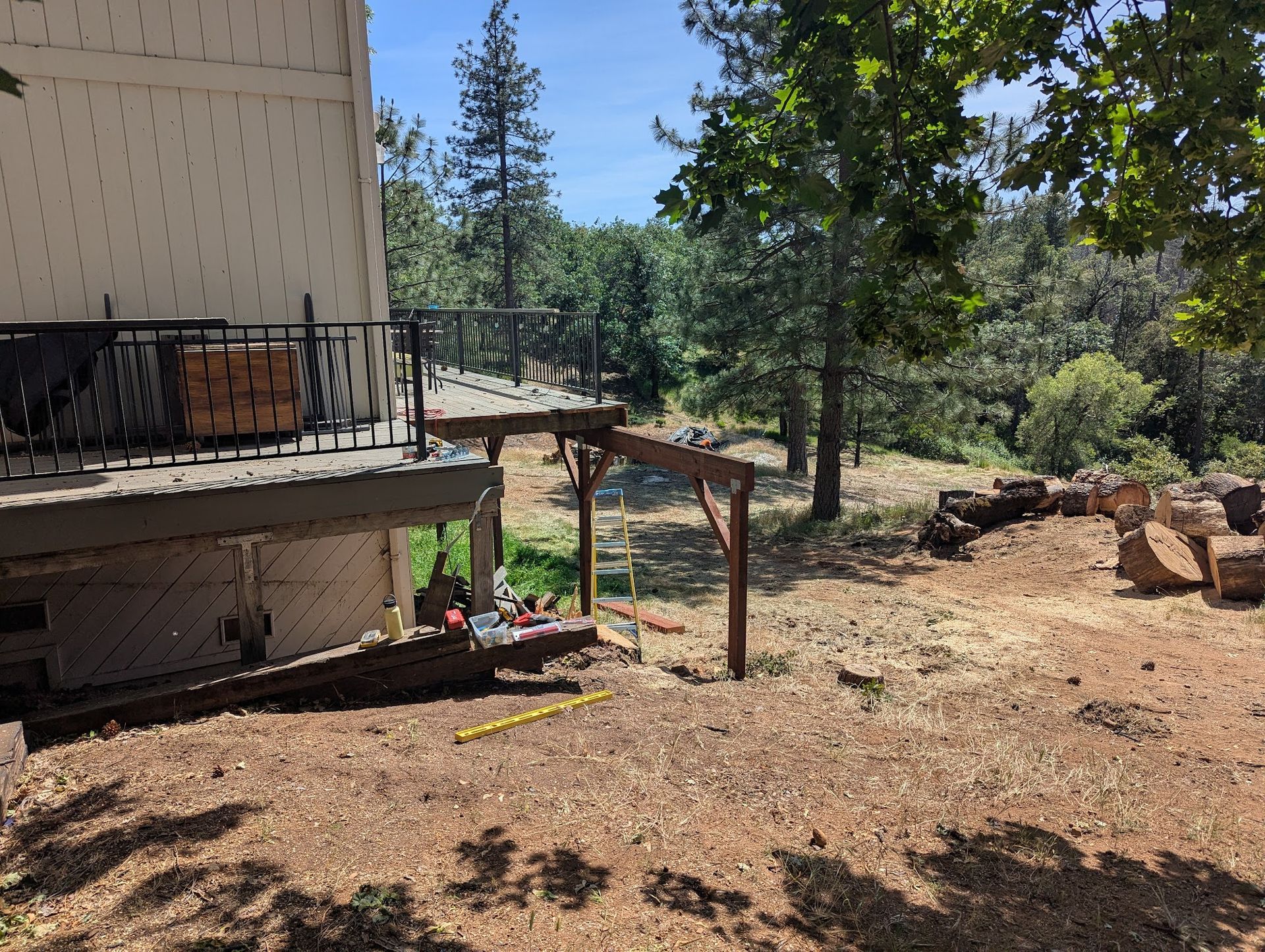 Backyard with a house deck, trees, dry ground, and a pergola near a wooded hillside.