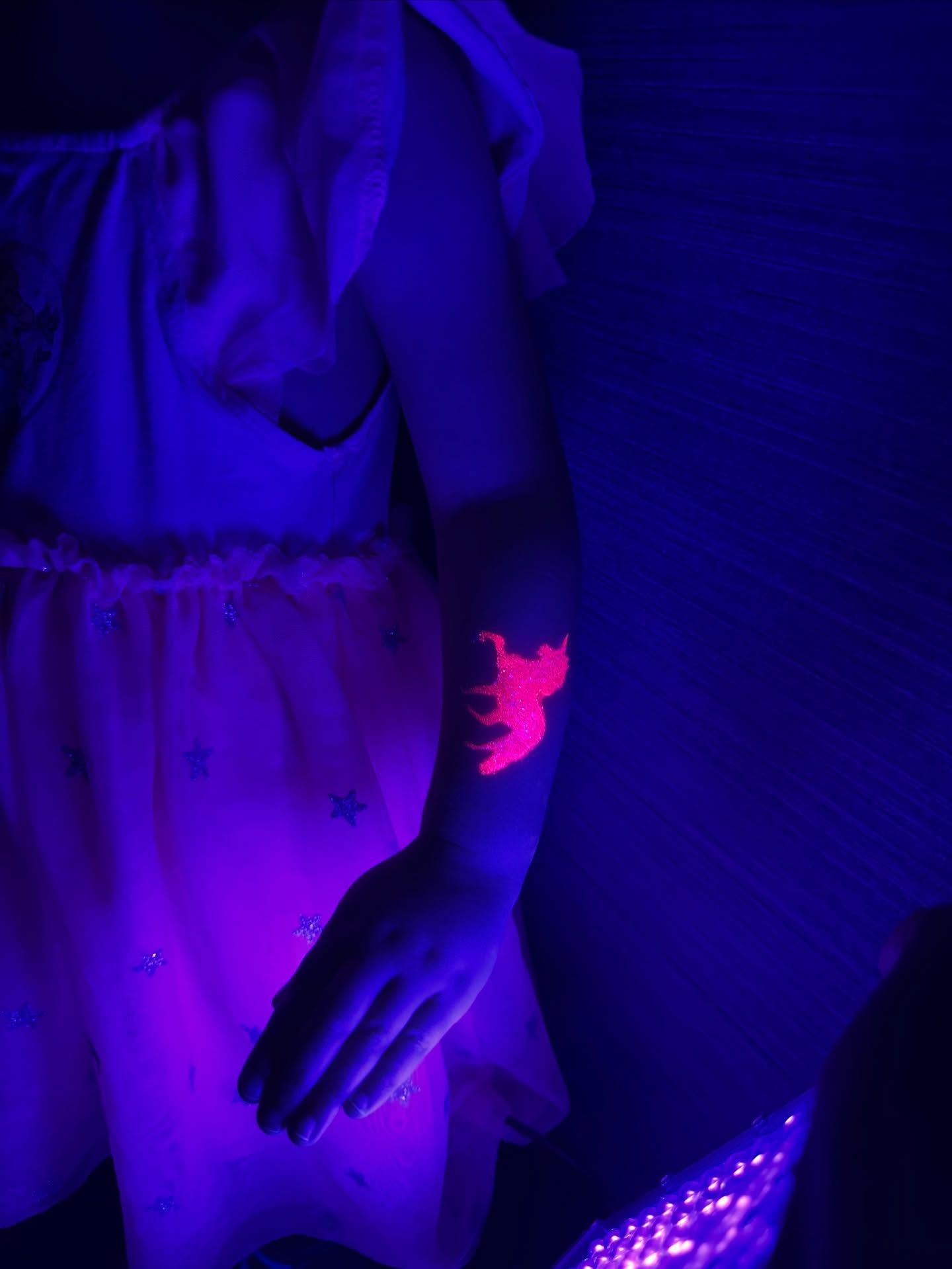 Person's arm with a glowing pink unicorn tattoo under a black light, arm in a pink dress, dark background.