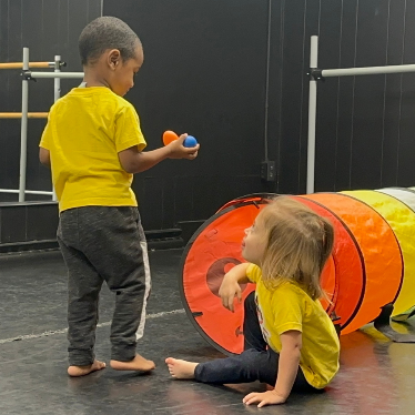 Two children in yellow shirts play near a tunnel. One holds a ball.