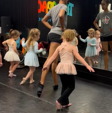 Young children in ballet class, practicing dance steps with instructors in a studio with a mirror.