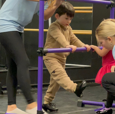 Boy in tan sweatsuit doing dance barre exercises, purple barre, teacher assisting.