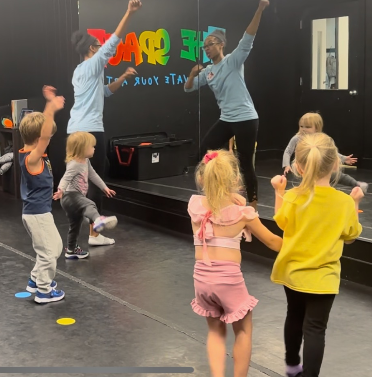 Children in a dance class follow instructors. They are raising their arms and dancing in a studio.