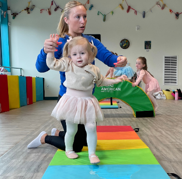 Woman assisting a toddler walking on a rainbow mat in a brightly lit room with other children.
