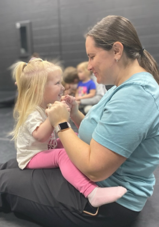 Woman sitting, smiling at toddler on her lap. Child holds woman's hands, giggling. Other children seated behind them.