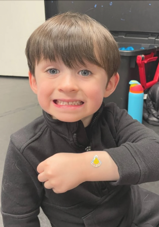 Young child smiles, showing a sticker on his hand. He is indoors, wearing a black jacket.