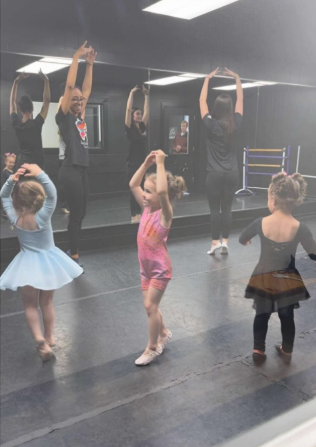 Ballet class with children and adults. Students stretch arms overhead in a mirrored studio.