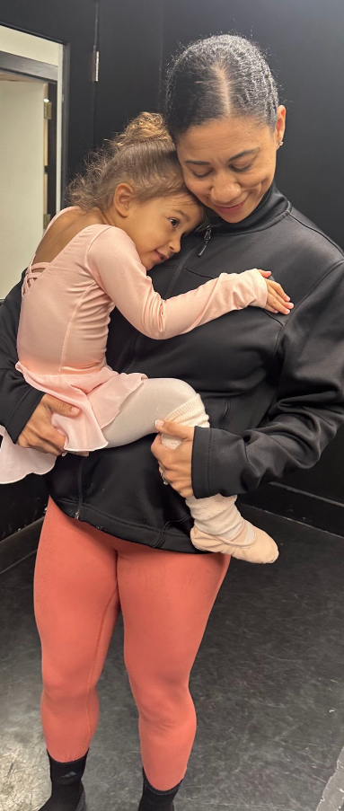 Woman holding a child dressed in a pink ballet outfit. The child rests their head on the woman's shoulder.