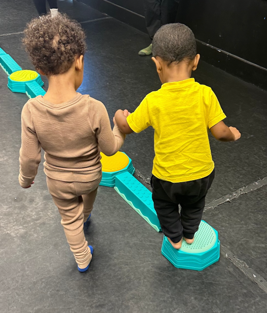 Two young children walking on a balance beam, holding hands. One wears brown, the other yellow.