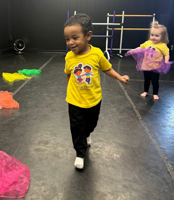 Boy in yellow shirt walks in a dance studio, another child in background.