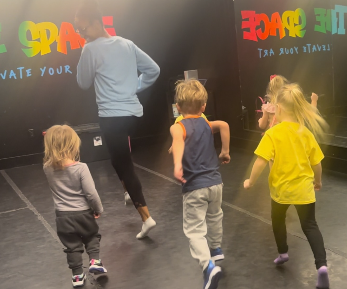 Children running in a dance studio, following a person in light blue; studio has colorful graffiti art.