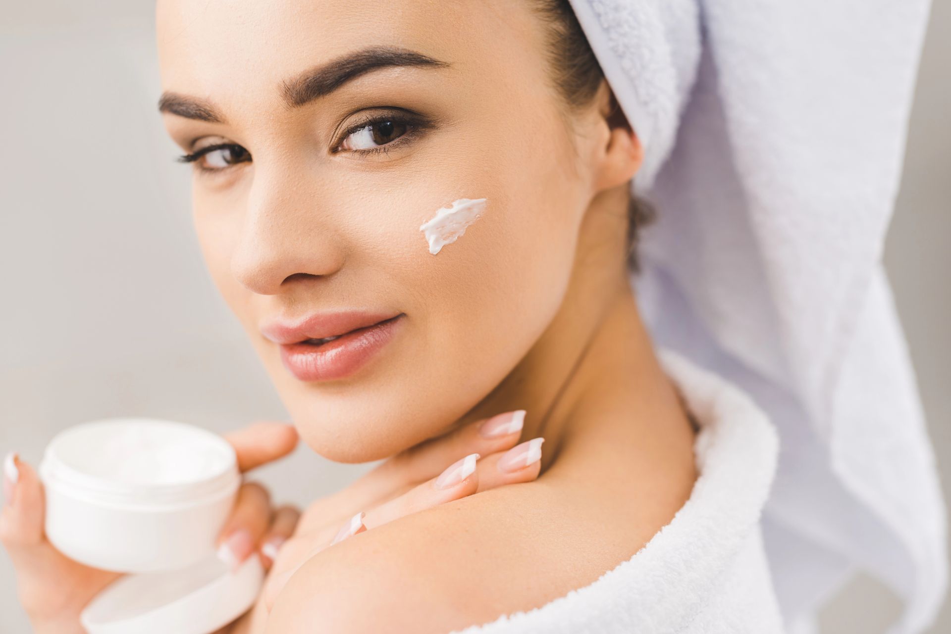 Mujer aplicándose crema en la cara y los hombros, envuelta en una toalla blanca.