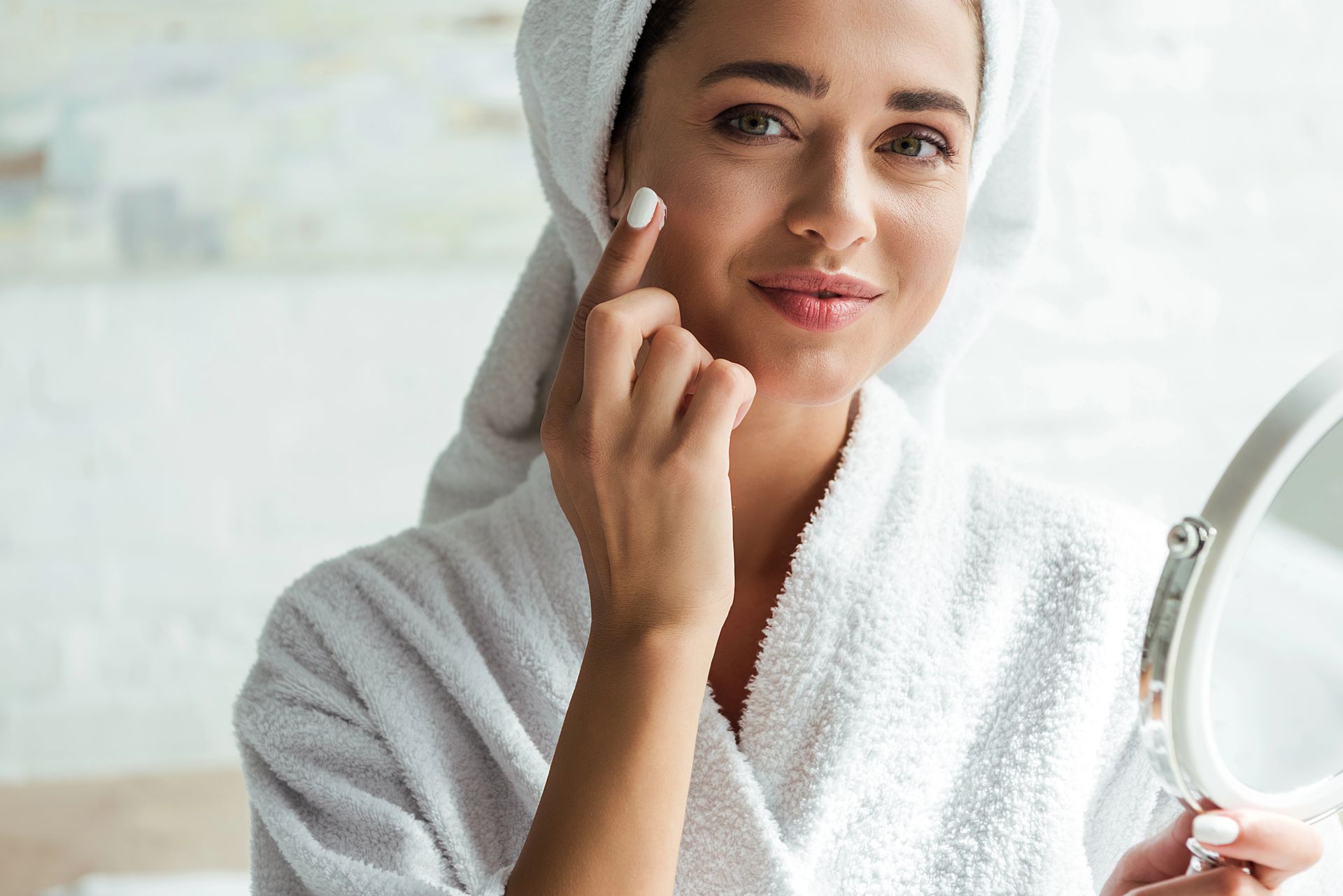 Mujer en bata de baño y toalla aplicándose productos para el cuidado de la piel, sosteniendo un espejo, sonriendo.