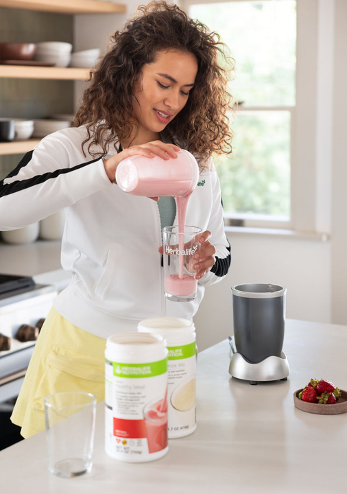 Mujer vertiendo un batido rosa de una licuadora en un vaso en la cocina. Ingredientes visibles.
