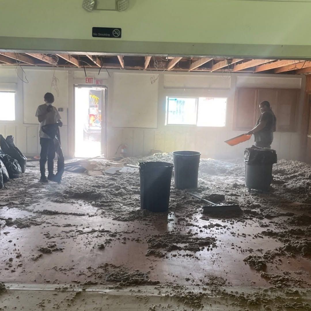 Two workers in a dusty, debris-covered room with exposed ceiling beams and an open doorway