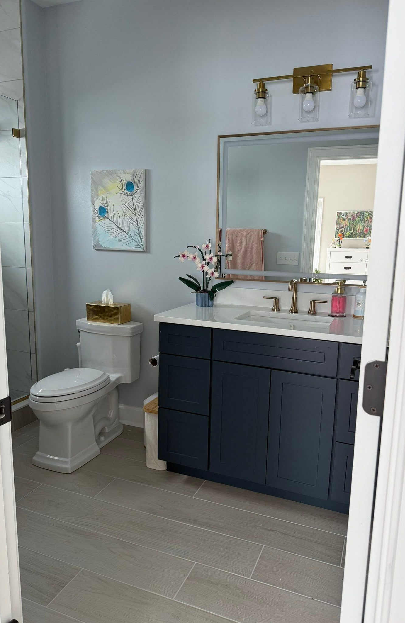 Compact bathroom with navy vanity, white sink, mirror, toilet, and brass light fixture on light gray walls.