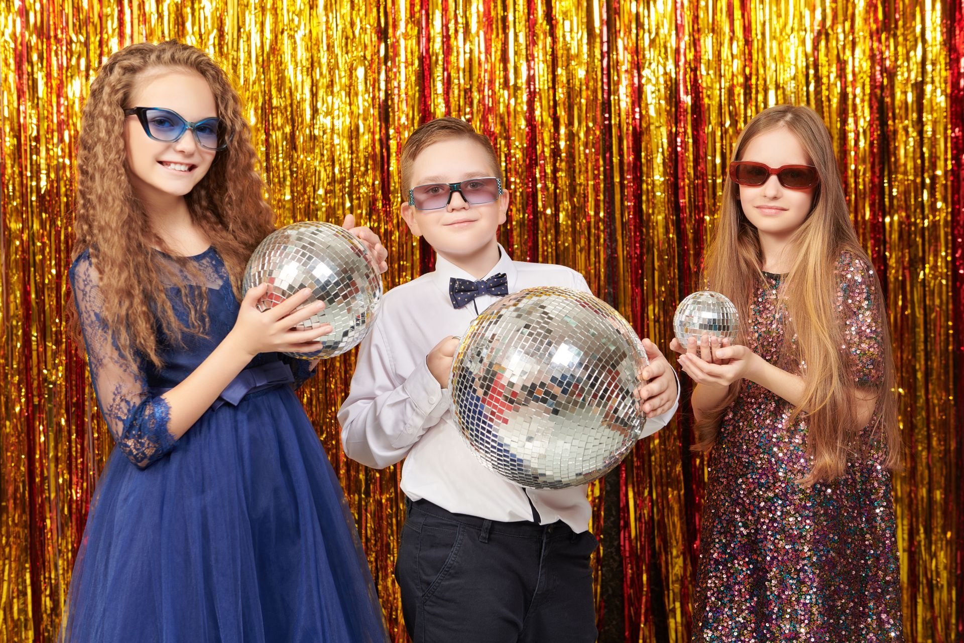 Three children with disco balls and sunglasses in front of a gold fringed backdrop.