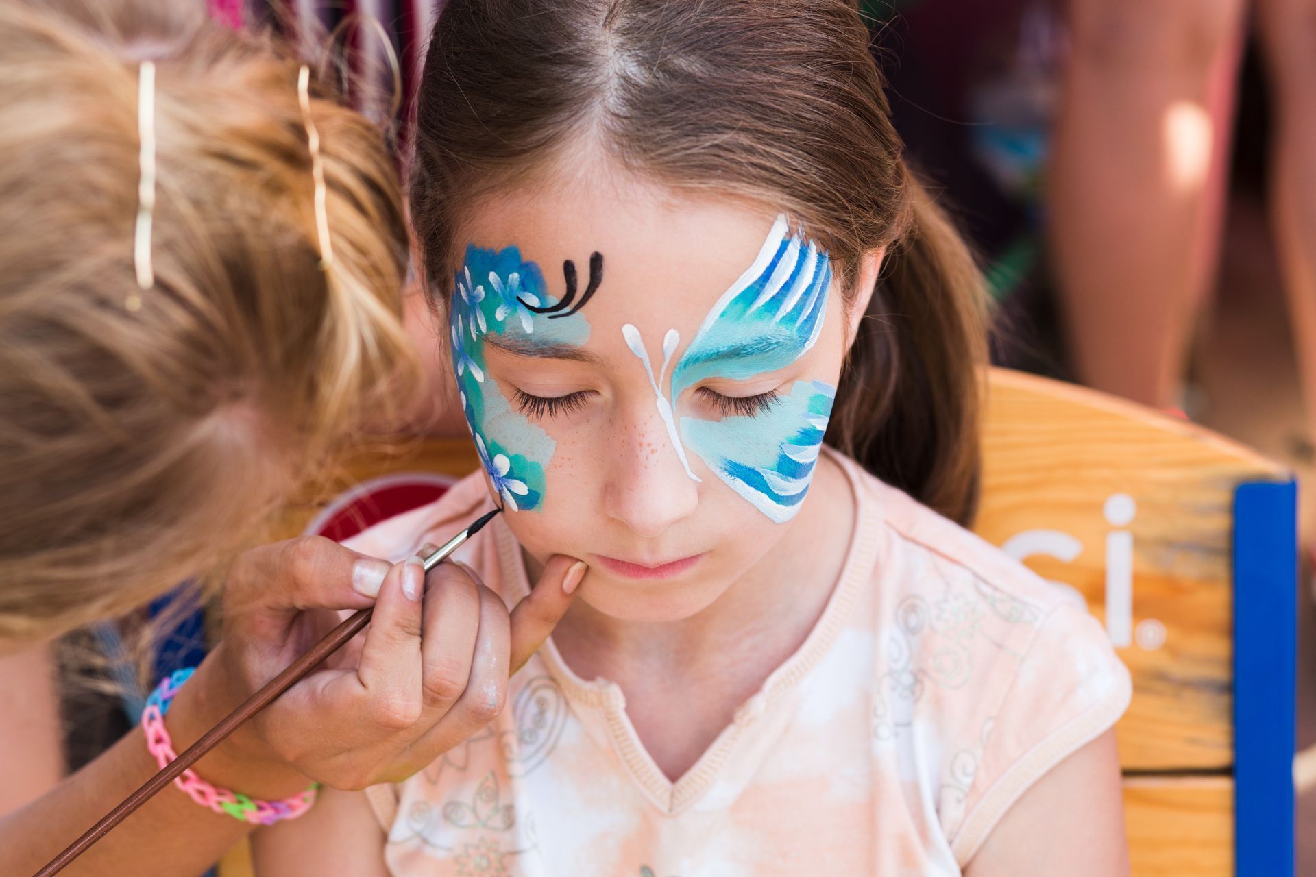A child with a blue and white butterfly face paint being applied by a person.