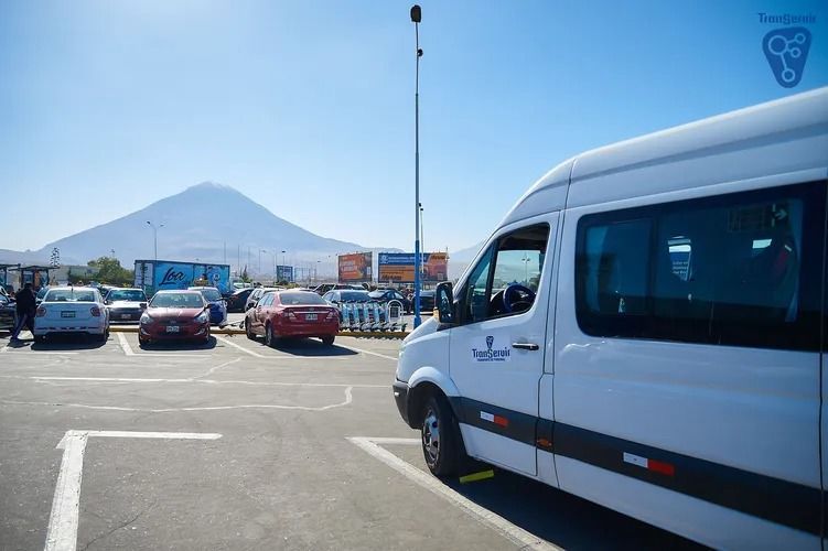 Aparcamiento con coches y una furgoneta blanca, montaña al fondo en un día soleado.