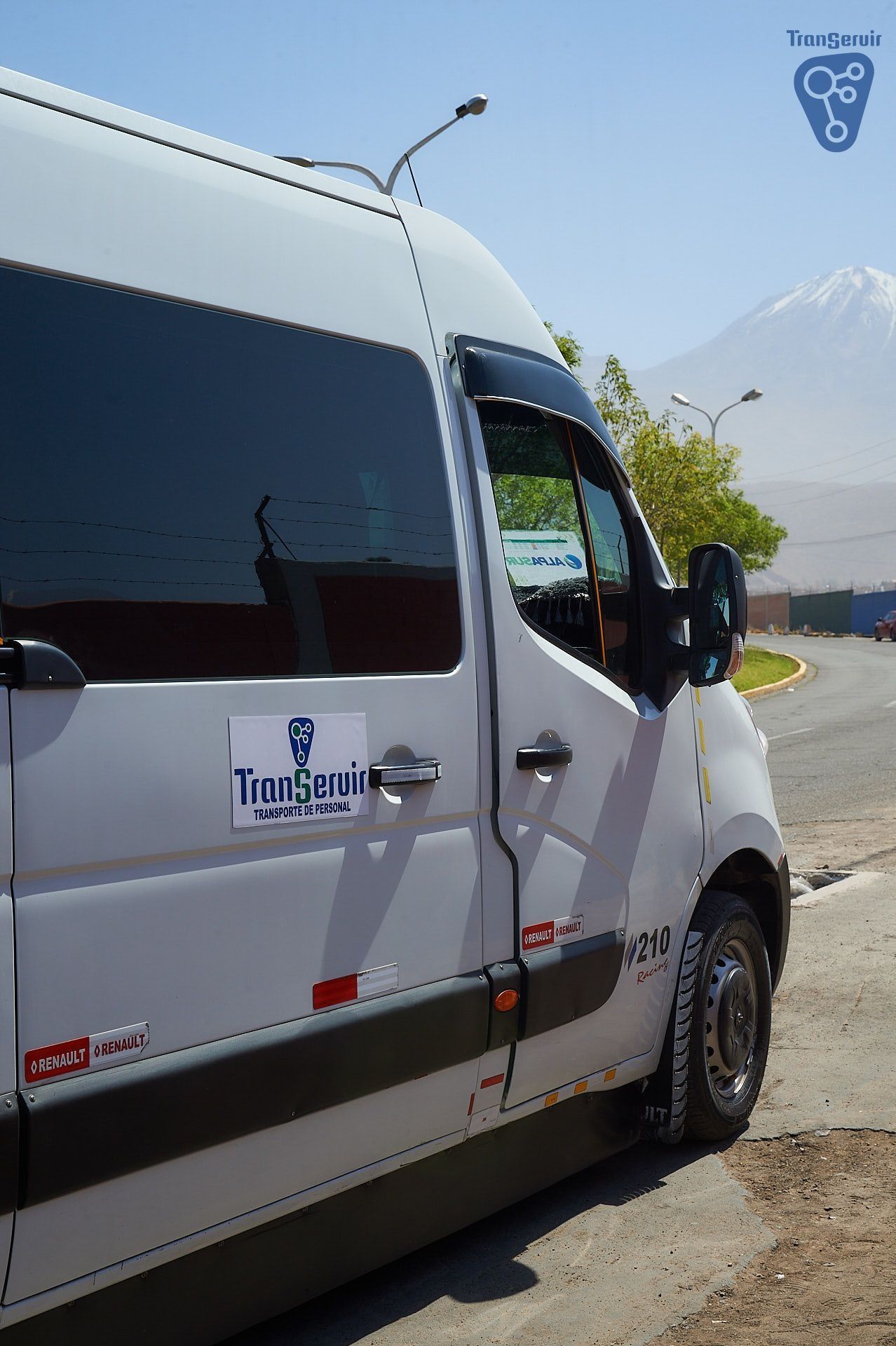 Furgoneta blanca aparcada al borde de la carretera con una montaña de fondo y un logotipo en la puerta.