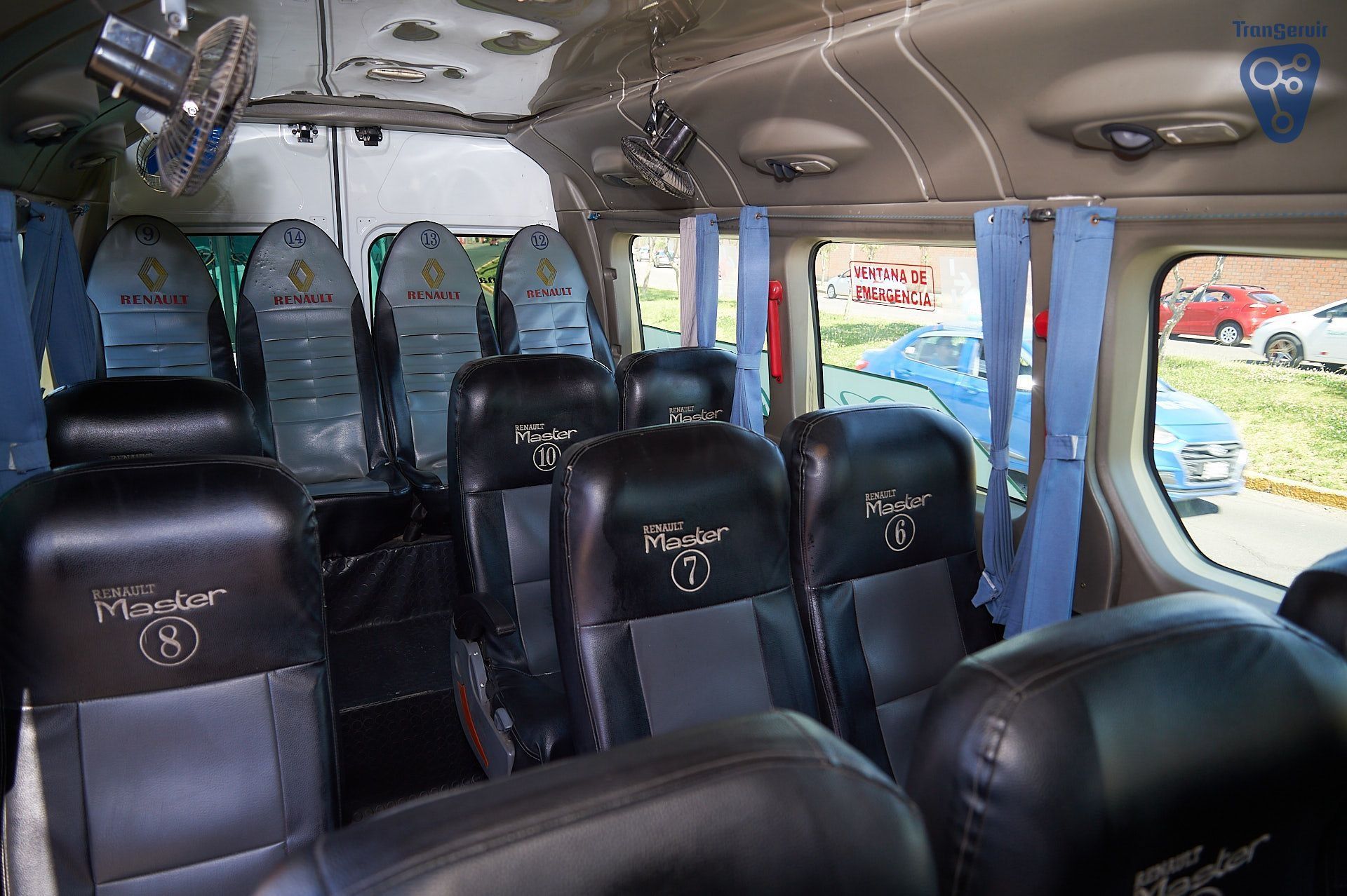 Interior de un minibús con asientos negros y cortinas azules, visto desde la parte trasera hacia el parabrisas delantero.