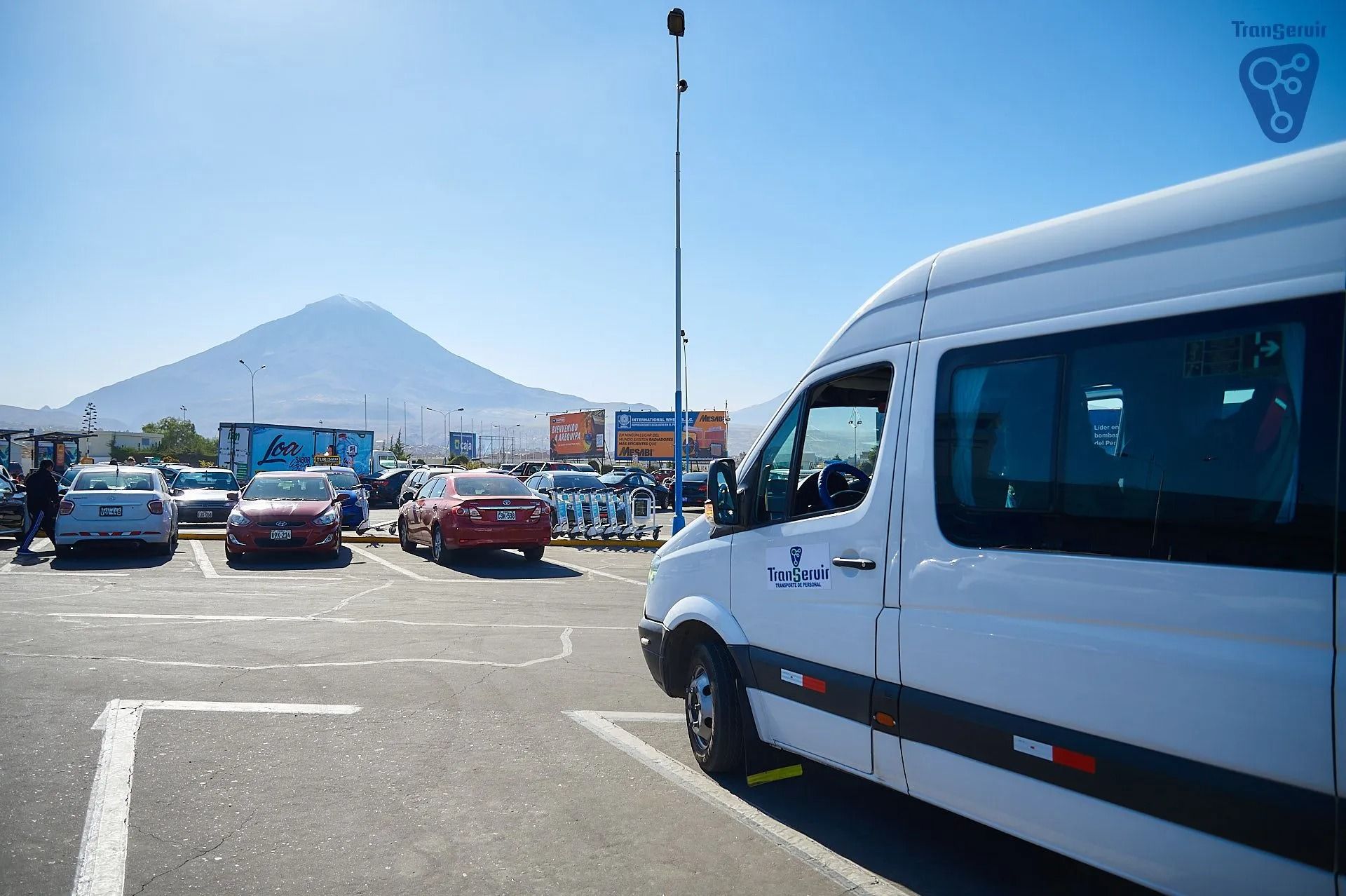 Tráfico en un estacionamiento con autos y una camioneta blanca, montañas al fondo bajo un cielo azul despejado.