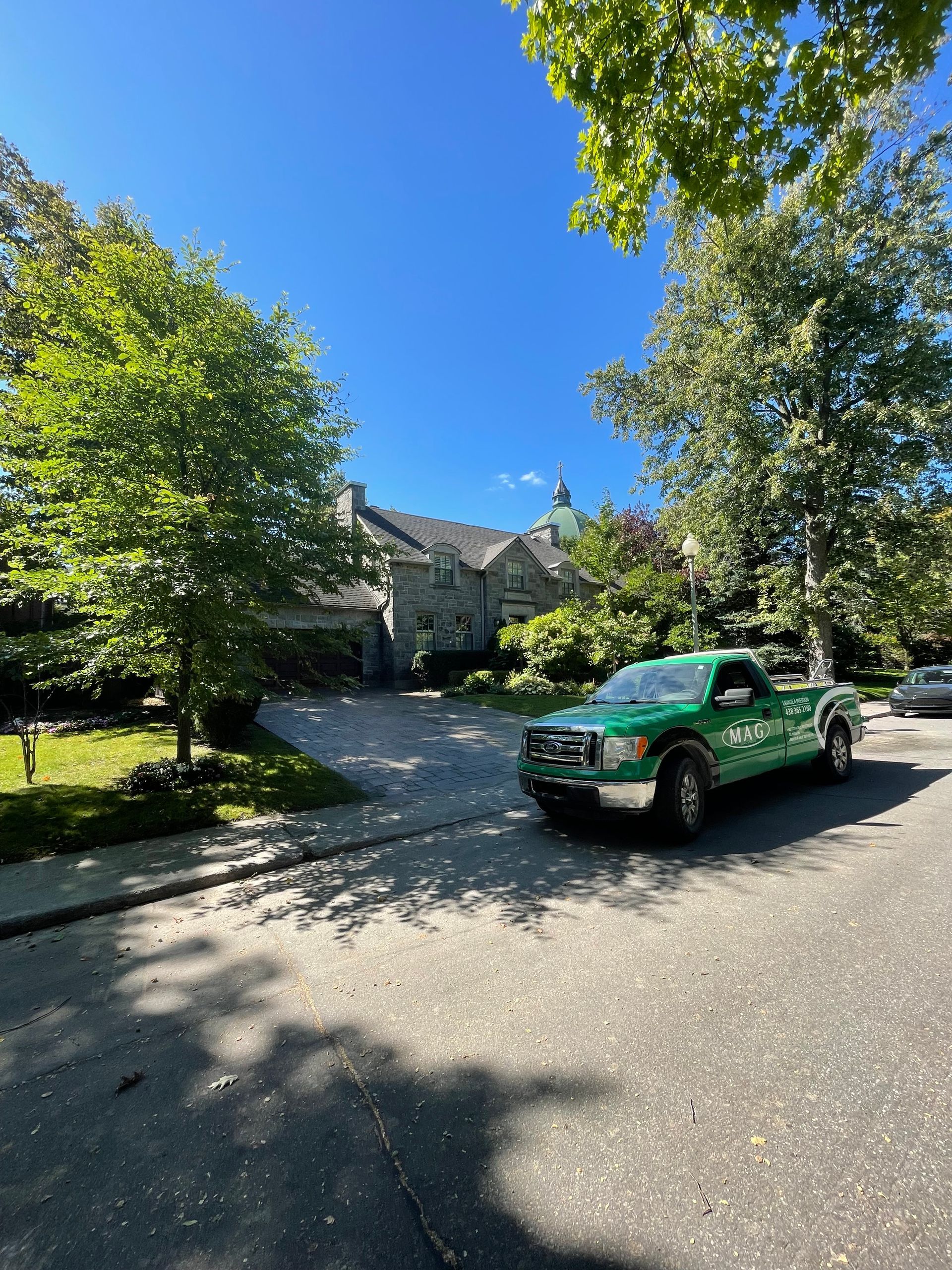 Camion vert garé dans la rue, devant une maison en pierre, avec des arbres et un ciel bleu.