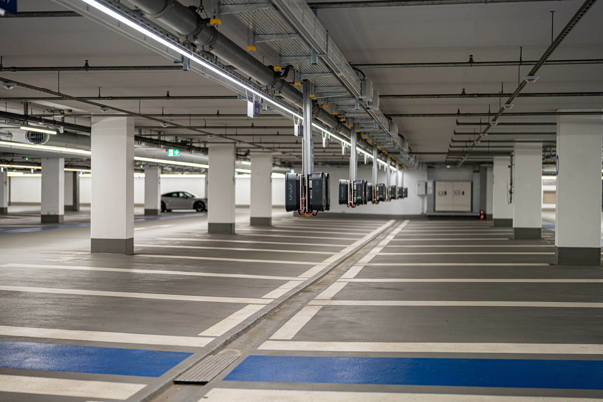 A spacious underground parking garage with white pillars, grey concrete floor, blue painted lines, and overhead lighting.