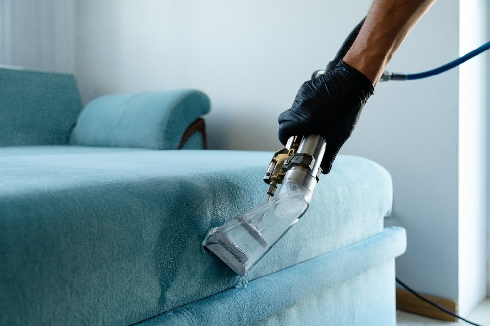A person wearing a black glove uses a cleaning machine to clean a teal sofa.